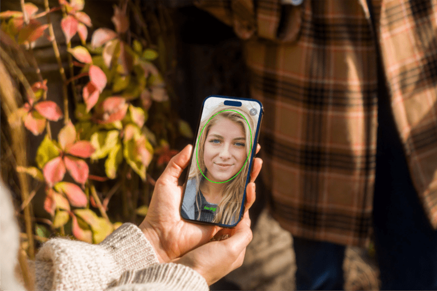 En person holder en smarttelefon med fokus på skjermen, som viser et bilde av en smilende ung kvinne med blondt hår. Det ser ut som om hun tar et bilde av seg selv gjennom en mobilapp.